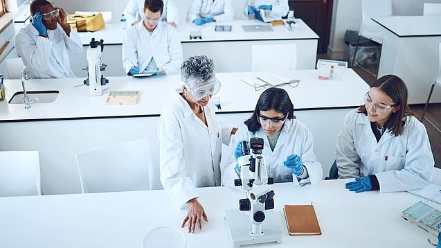 Gruppe Studierender und eine Dozentin in einem Chemiehörsaal vor Mikroskopen