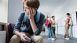 Ein Junge sitzt mit einem Handy in der Hand auf einem Schulflur und schaut traurig aus dem Fenster.