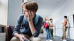 Ein Junge sitzt mit einem Handy in der Hand auf einem Schulflur und schaut traurig aus dem Fenster.