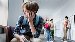 Ein Junge sitzt mit einem Handy in der Hand auf einem Schulflur und schaut traurig aus dem Fenster.