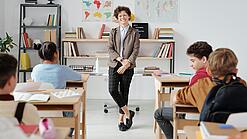 Junge Lehrerin steht vor einer Grundschulklasse und lächelt in die Kamera.