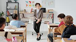 Junge Lehrerin steht vor einer Grundschulklasse und lächelt in die Kamera.