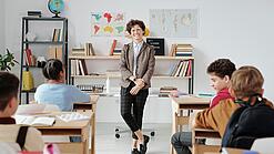 Junge Lehrerin steht vor einer Grundschulklasse und lächelt in die Kamera.