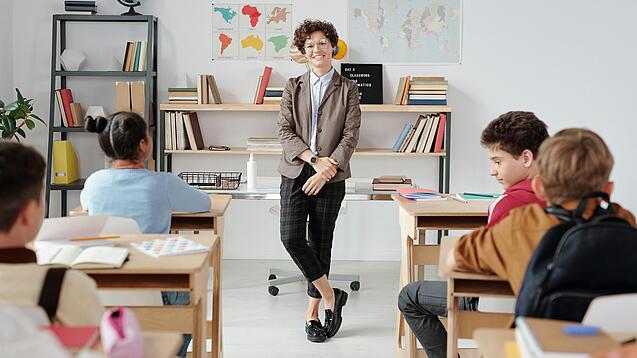 Junge Lehrerin steht vor einer Grundschulklasse und lächelt in die Kamera.
