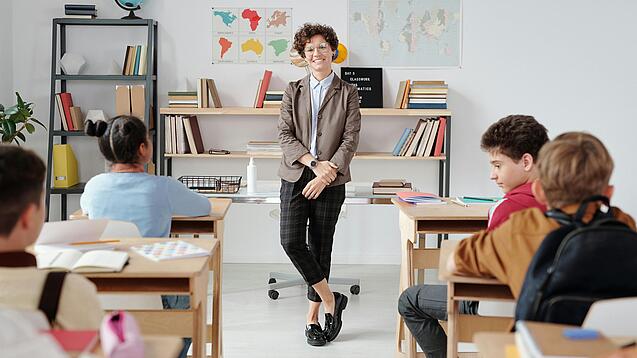Junge Lehrerin steht vor einer Grundschulklasse und lächelt in die Kamera.