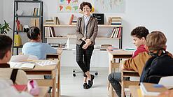 Junge Lehrerin steht vor einer Grundschulklasse und lächelt in die Kamera.