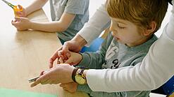 Ein Junge im Kindergartenalter wird beim Schneiden mit einer Schere unterstützt.