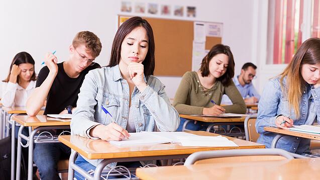 Mehrere Schülerinnen und Schüler sitzen an ihren Tischen in einem Klassenzimmer und arbeiten konzentriert in ihren Heften.
