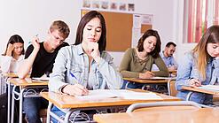 Mehrere Schülerinnen und Schüler sitzen an ihren Tischen in einem Klassenzimmer und arbeiten konzentriert in ihren Heften.