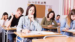 Mehrere Schülerinnen und Schüler sitzen an ihren Tischen in einem Klassenzimmer und arbeiten konzentriert in ihren Heften.