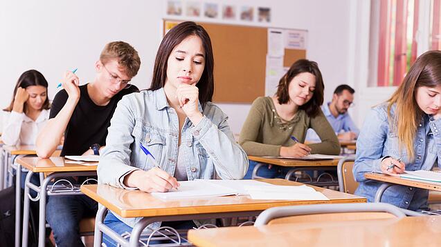 Mehrere Schülerinnen und Schüler sitzen an ihren Tischen in einem Klassenzimmer und arbeiten konzentriert in ihren Heften.