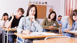 Mehrere Schülerinnen und Schüler sitzen an ihren Tischen in einem Klassenzimmer und arbeiten konzentriert in ihren Heften.