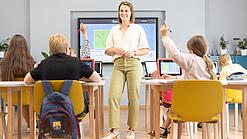 Eine Lehrerin steht in einem Klassenzimmer, Schülerinnen und Schüler sitzen an Tischen.