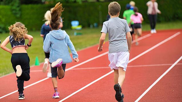 Drei Kinder laufen auf einem Sportplatz über eine Laufbahn.