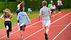 Drei Kinder laufen auf einem Sportplatz über eine Laufbahn.