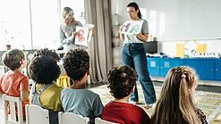 Zwei Erzieherinnen stehen vor einer Gruppe Kita-Kinder und erklärt ihnen etwas.