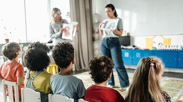 Zwei Erzieherinnen stehen vor einer Gruppe Kita-Kinder und erklärt ihnen etwas.