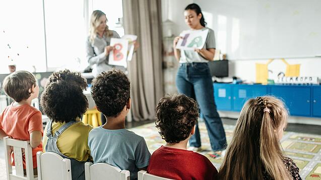Zwei Erzieherinnen stehen vor einer Gruppe Kita-Kinder und erklärt ihnen etwas.