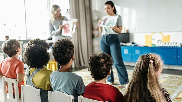 Zwei Erzieherinnen stehen vor einer Gruppe Kita-Kinder und erklärt ihnen etwas.
