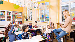Grundschulkinder sitzen in einem Klassenraum und hören einer Lehrerin zu.