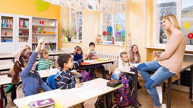 Grundschulkinder sitzen in einem Klassenraum und hören einer Lehrerin zu.