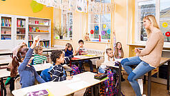 Grundschulkinder sitzen in einem Klassenraum und hören einer Lehrerin zu.