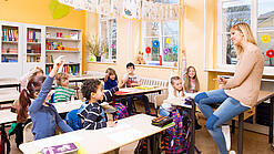 Grundschulkinder sitzen in einem Klassenraum und hören einer Lehrerin zu.