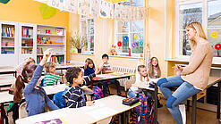 Grundschulkinder sitzen in einem Klassenraum und hören einer Lehrerin zu.