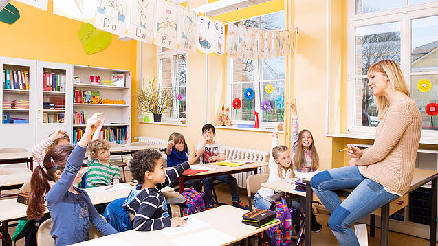 Grundschulkinder sitzen in einem Klassenraum und hören einer Lehrerin zu.