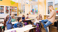 Grundschulkinder sitzen in einem Klassenraum und hören einer Lehrerin zu.