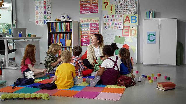 Gruppe Grundschulkinder und eine Lehrerin sitzen am Boden in einem Kreis.