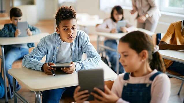Schulkinder sitzen in einem Klassenraum und zeigen sich gegenseitig etwas auf einem Tablet.