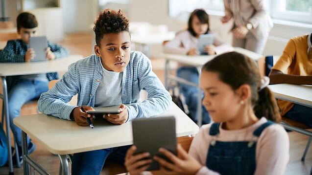 Schulkinder sitzen in einem Klassenraum und zeigen sich gegenseitig etwas auf einem Tablet.