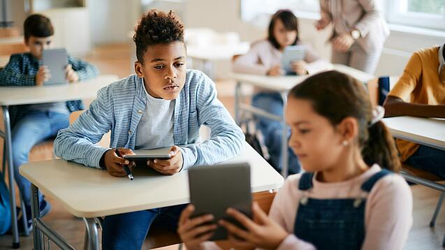 Schulkinder sitzen in einem Klassenraum und zeigen sich gegenseitig etwas auf einem Tablet.