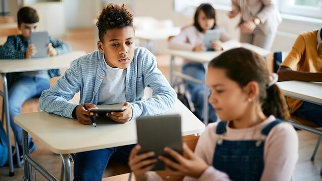Schulkinder sitzen in einem Klassenraum und zeigen sich gegenseitig etwas auf einem Tablet.