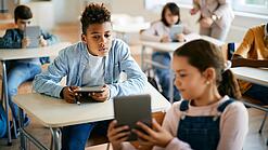 Schulkinder sitzen in einem Klassenraum und zeigen sich gegenseitig etwas auf einem Tablet.