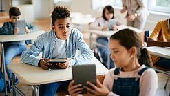 Schulkinder sitzen in einem Klassenraum und zeigen sich gegenseitig etwas auf einem Tablet.