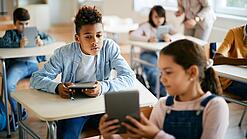 Schulkinder sitzen in einem Klassenraum und zeigen sich gegenseitig etwas auf einem Tablet.
