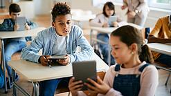 Schulkinder sitzen in einem Klassenraum und zeigen sich gegenseitig etwas auf einem Tablet.