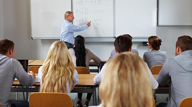 Ein Lehrer steht in einer Klasse an einer Tafel und erklärt etwas.