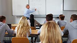 Ein Lehrer steht in einer Klasse an einer Tafel und erklärt etwas.