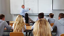 Ein Lehrer steht in einer Klasse an einer Tafel und erklärt etwas.