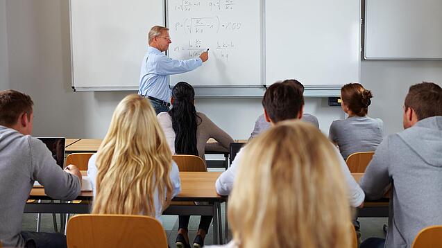 Ein Lehrer steht in einer Klasse an einer Tafel und erklärt etwas.