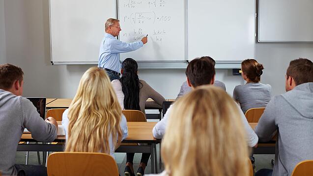 Ein Lehrer steht in einer Klasse an einer Tafel und erklärt etwas.