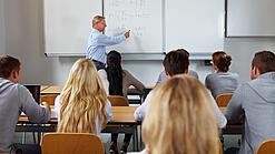Ein Lehrer steht in einer Klasse an einer Tafel und erklärt etwas.