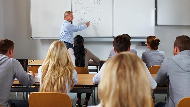 Ein Lehrer steht in einer Klasse an einer Tafel und erklärt etwas.
