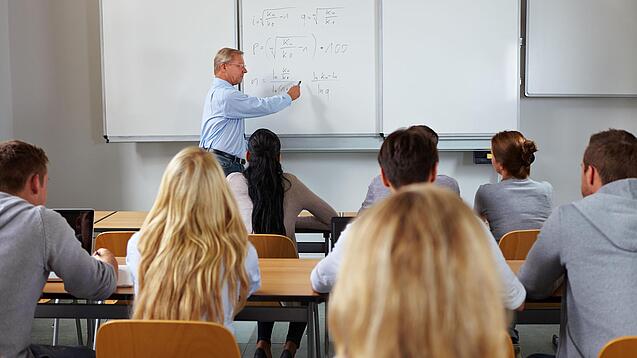 Ein Lehrer steht in einer Klasse an einer Tafel und erklärt etwas.