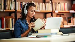 Junge Frau mit Kopfhören sitzt in einer Bibliothek vor einem Laptop und blättert in einem Buch.