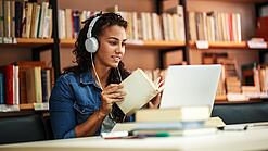 Junge Frau mit Kopfhören sitzt in einer Bibliothek vor einem Laptop und blättert in einem Buch.