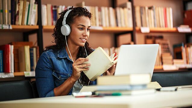 Junge Frau mit Kopfhören sitzt in einer Bibliothek vor einem Laptop und blättert in einem Buch.